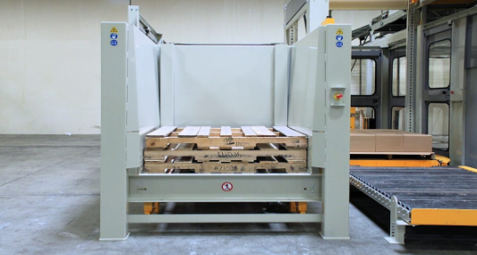 Empty wooden pallets sit inside an automated pallet dispenser beside a roller conveyor on a manufacturing floor.